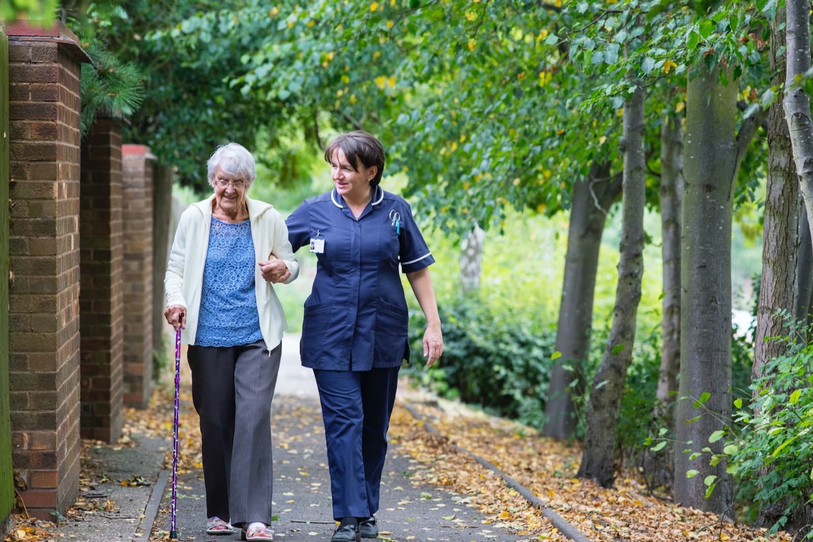 A carer walking arm in arm with an older woman along a leafy path